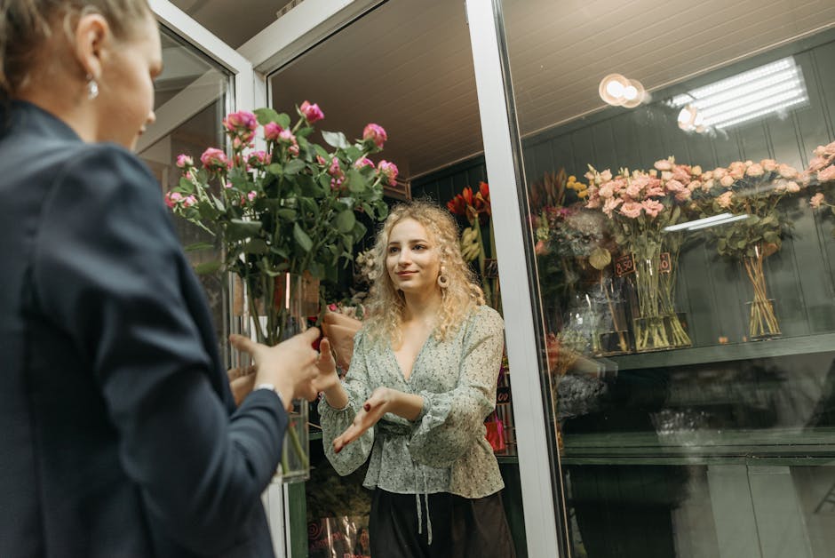 A young woman with curly blonde hair, wearing a light patterned blouse, stands inside a flower shop with glass display cases filled with pink and peach roses in large vases, as she receives a bouquet 