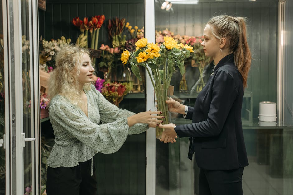 A woman with curly blonde hair wearing a light-colored blouse with a floral pattern is receiving a tall floral bouquet featuring bright yellow sunflowers and green stems, wrapped in clear cellophane, 