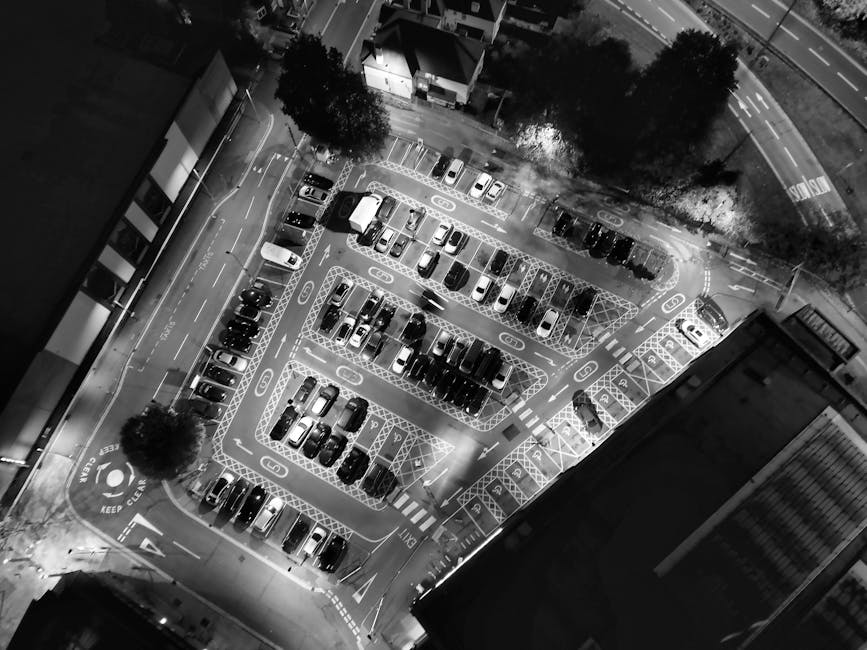 A black-and-white aerial photograph of a parking lot illuminated by bright overhead lighting, showing multiple rows of parked cars, clearly marked parking spaces, and surrounding trees. The image capt
