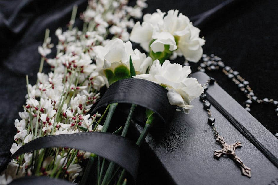 A close-up of a bouquet containing white carnations and smaller white flowers with pinkish tips, arranged with green stems, resting on a black fabric surface. The bouquet is partially wrapped with bla