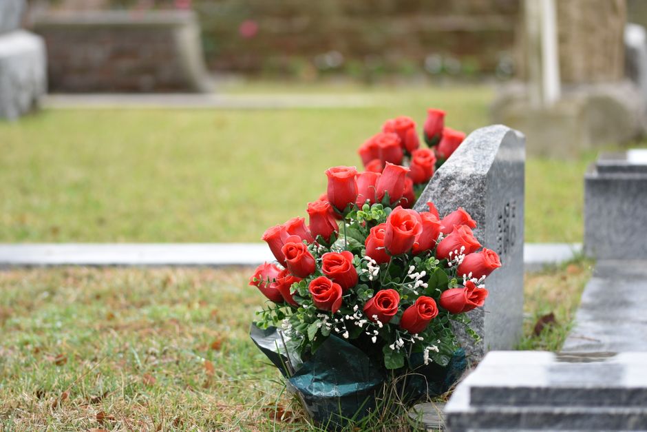 A bouquet arrangement of fresh red roses, with tightly closed and open petals, is placed against a grey stone gravestone at a cemetery. The bouquet is wrapped in dark green paper and tied with a ribbo