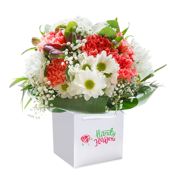 Framed by a spotless white backdrop, a striking floral arrangement stands proudly in a square white gift bag-an image that embodies the welcoming spirit of Emerson Park. This bouquet is a masterful blend of soft coral and radiant red carnations, each petal ruffled and abundant, exuding a sense of gentle luxury. Bright white daisies with zesty yellow-green centers are interspersed throughout, each one a cheerful nod to the sunny open spaces of Emerson Park's parks and lanes. Airy, white clouds of baby's breath drift between the blooms, delivering a delicate, almost dreamy texture, while generous deep green foliage gathers around the base. Glossy, broad leaves fan out, providing a rich verdant foundation that hints at the well-tended private gardens that dot the local neighbourhood. On the left side, a few unopened purple-tinged buds add intrigue and natural depth, offering the promise of future blossoms-much like the evolving seasons in the community. The flowers are carefully shaped into a harmonious, rounded form, their freshness and vitality evident even from afar. The crisp white gift bag is topped with strong white ribbon handles, reflecting the same care and attention given to each element in this arrangement. Perhaps chosen for a milestone birthday, an anniversary in Emerson Park, or as a thoughtful gesture for a friend on Upminster Road, this bouquet typifies the elegance and heartfelt artistry found at Emerson Park Florist.